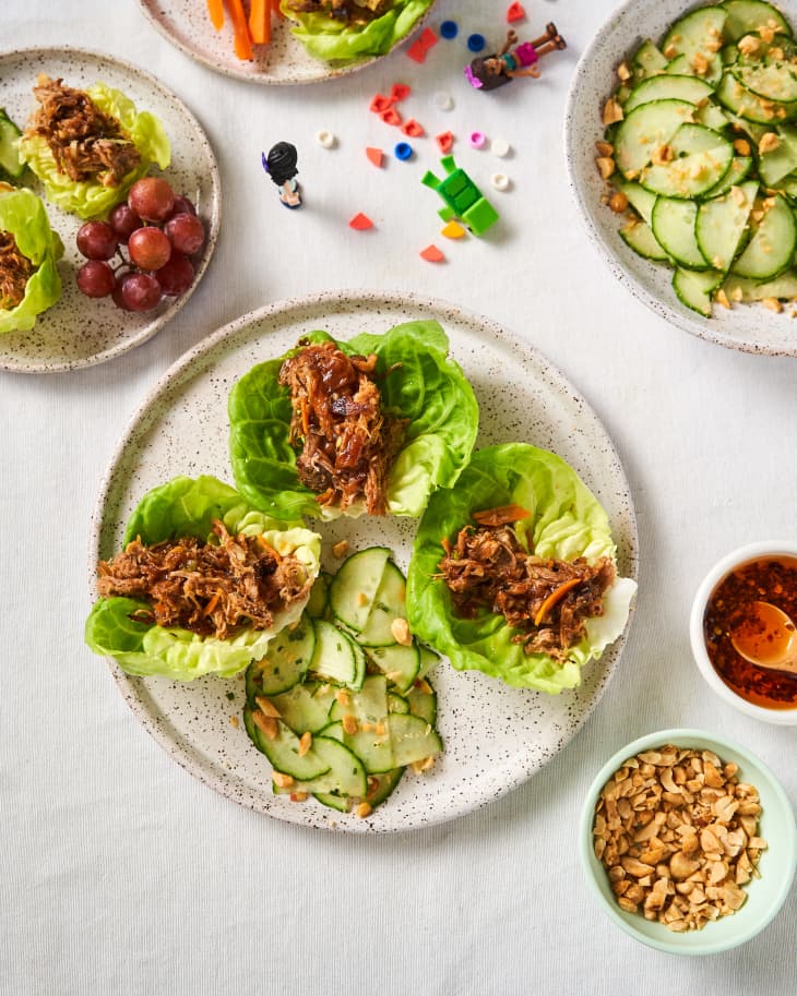 Speckled ceramic plate filled with shedded pork on top of Boston lettuce and pickles on the side. Smaller, colorful plates surround with pickles, grapes, chili sauce, peanuts and toy solider scattered on the table