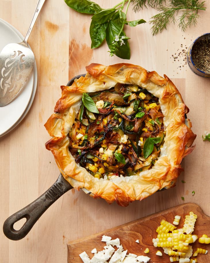 Vegetable phyllo pie in skillet surround by serving knife, fresh herbs, pepper and fresh corn cut off cob and shaved parmesan cheese