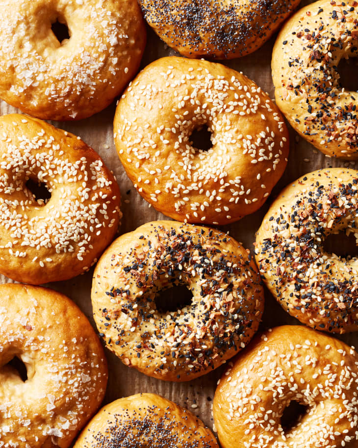 Ten bagels sit on a counter. Each bagel is topped with different seasonings like, everything seasoning, poppyseeds, sesame seeds, and salt.