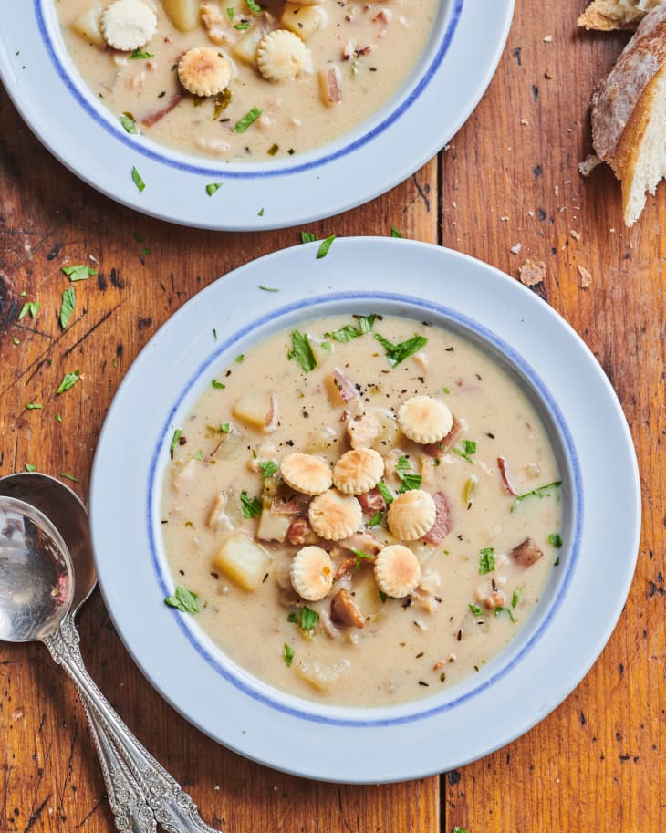 Two bowls of New England clam chowder topped with oyster crackers; soup spoons and bread on the side.