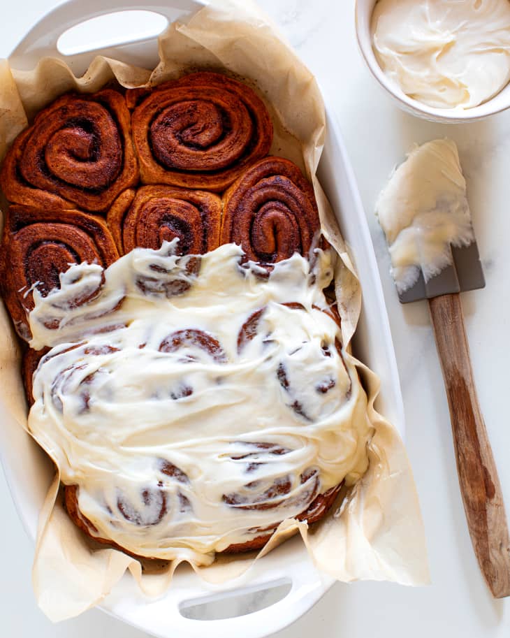 cinnamon rolls sit in a white baking tray half covered in icing with rubber spatula and tin of icing near it