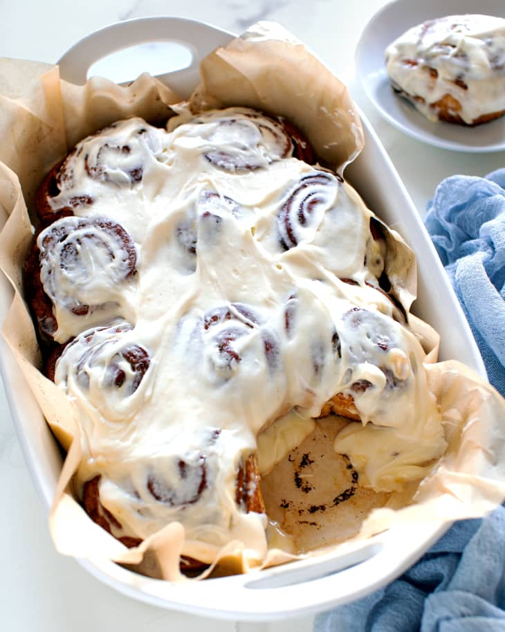 cinnamon rolls are in a white baking tin on top of parchment paper with one on a plate near it fully iced