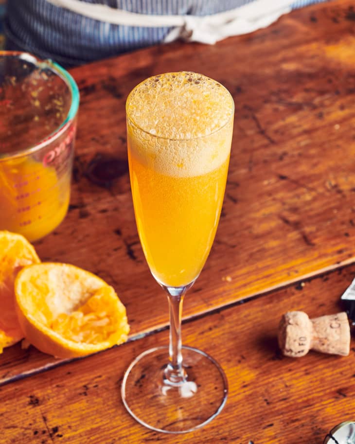 One mimosa sits on a wooden table with tangerine skins and a glass of tangerine juice behind it.
