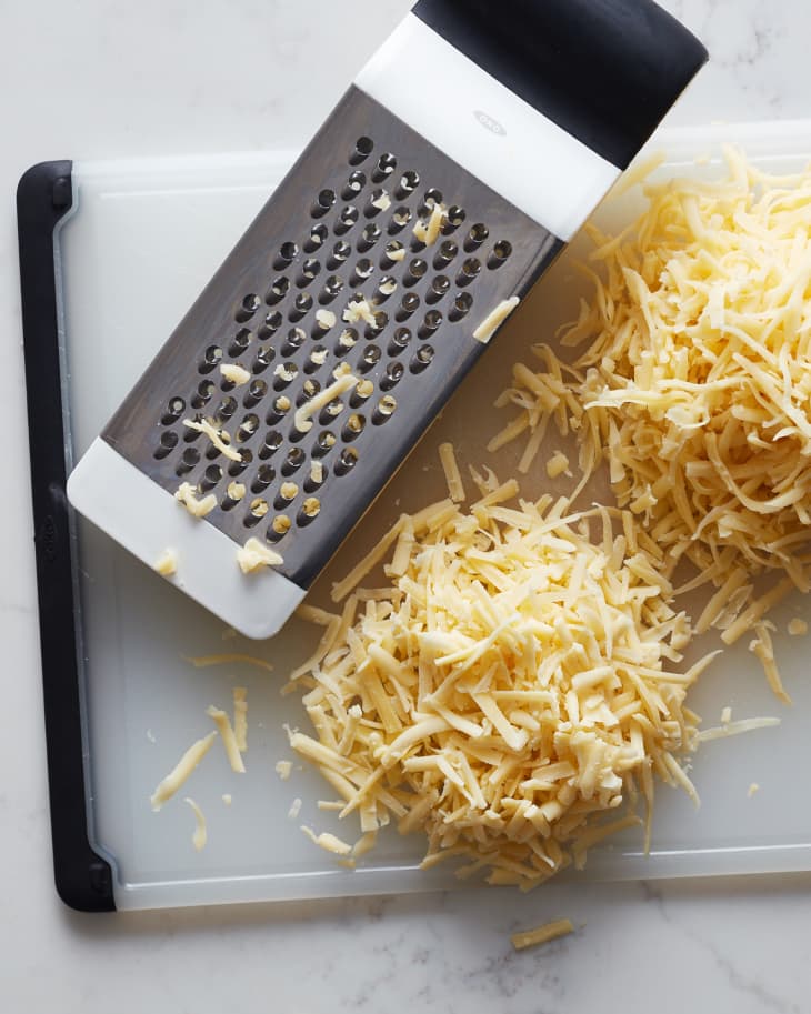 Cheese grater and piles of grated cheese on white cutting board