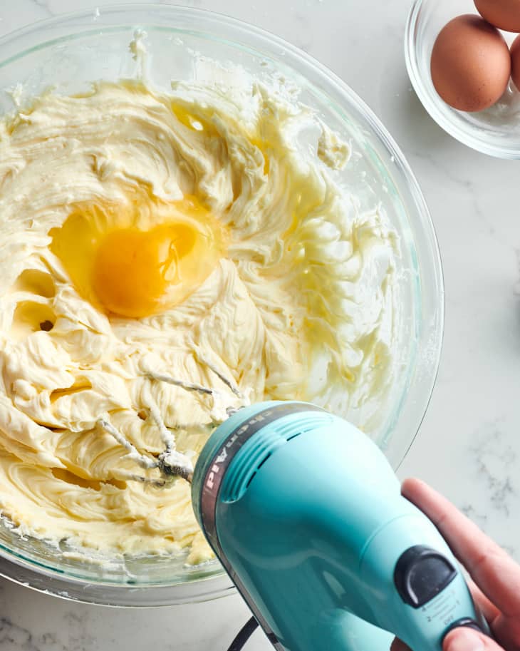 Overhead photo of someone mixing egg into batter with a hand mixer. Extra eggs in bowl in the background