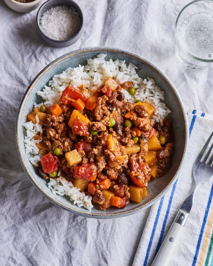 picadillo sits in a bowl over rice next to a glass and a fork