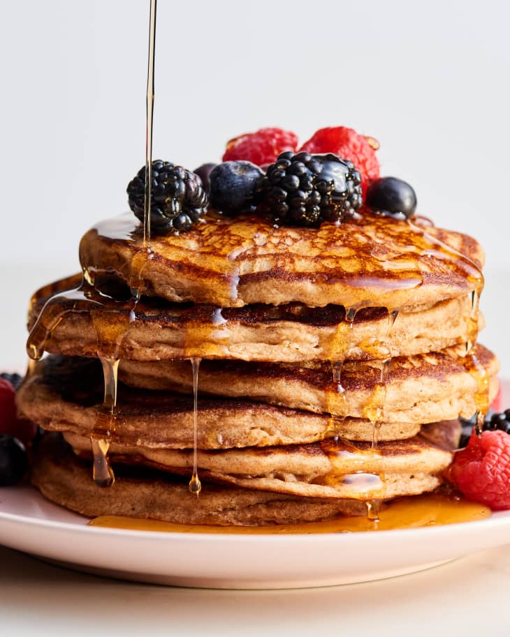 stack of whole wheat pancakes, topped with berries, being drizzled with maple syrup