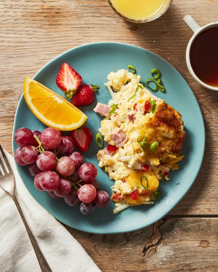 Aqua colored plate with one serving of easy breakfast casserole, with a side of red grapes, orange, strawberry. Coffee and orange juice in top right, fork and napkin in bottom left