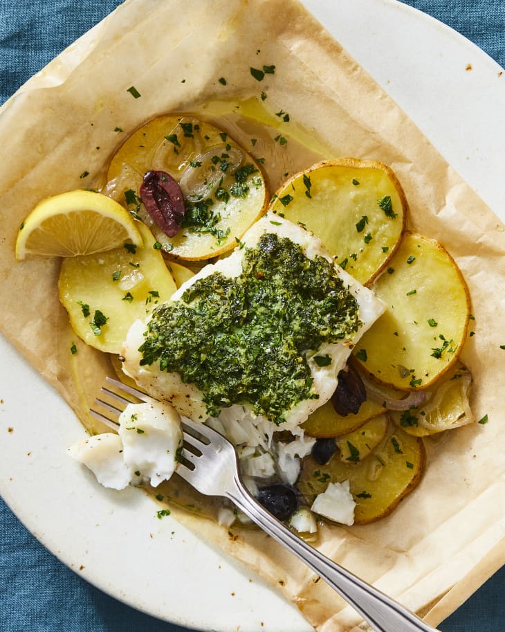 Filet of cod that's been baked with butter, herbs, potatoes. on parchment on a plate. Fork has cut out a bite