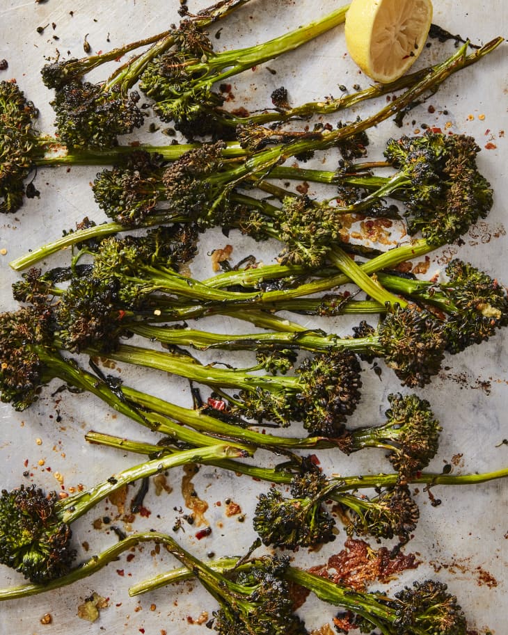 Overhead shot of roasted broccolini on sheet pan. Seasoned with red chili flakes, half a lemon in top of photo