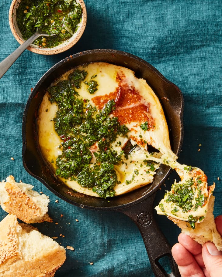 Overhead photo of small skillet with provolone that's been grilled, topped with chimichurri sauce. Someone is dipping in some crusty bread and there is a cheese pull. Small bowl of extra chimichurri in the upper left corner