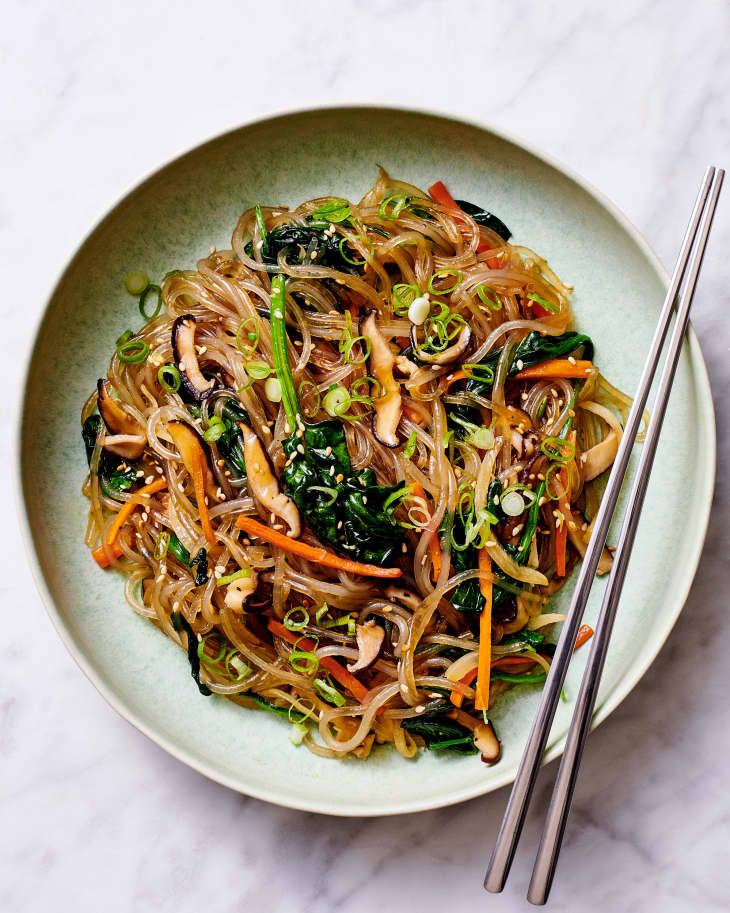 Japchae, or Korean sweet potato noodle stir-fried with carrots, spinach, and mushrooms, and garnished with green onions and sesame seeds, on a plate with chopsticks