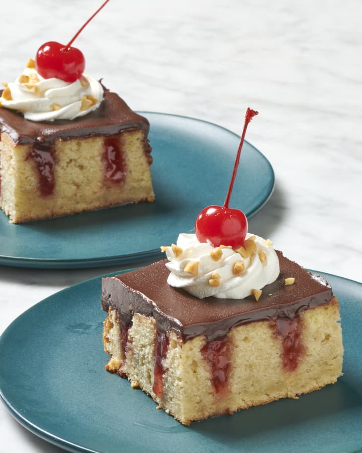 A piece of banana split poke cake with a cherry on top on a dark teal plate with another plate with a piece in the background