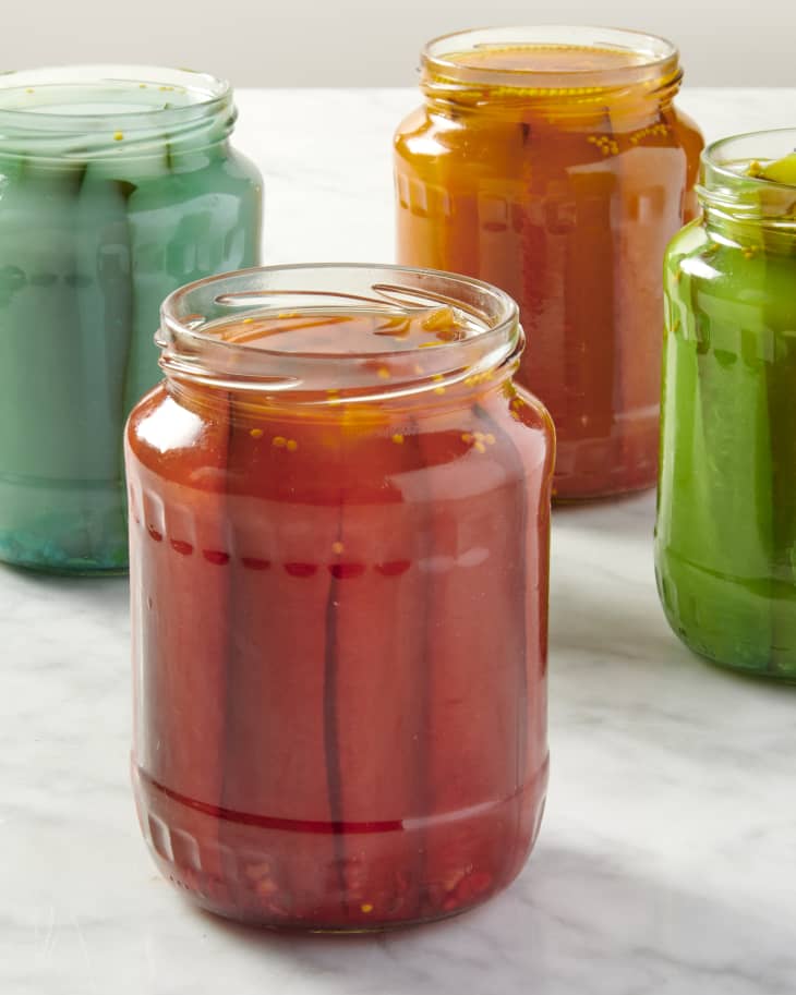 a group of four jars of pickles infused with blue, red, orange, and green Kool Aid on a marble countertop