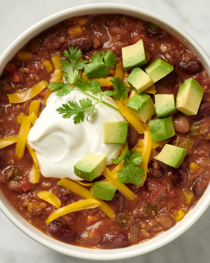 photo of a bowl of chili topped with shredded cheddar, avocado, parsley, and sour cream on a marble surface