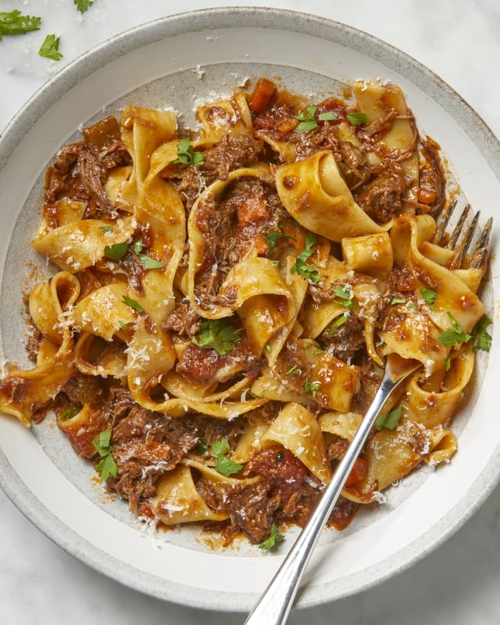 Overhead view of beef ragu on a grey and beige striped plate, topped with cheese and herbs, and a fork on the right side of the plate.