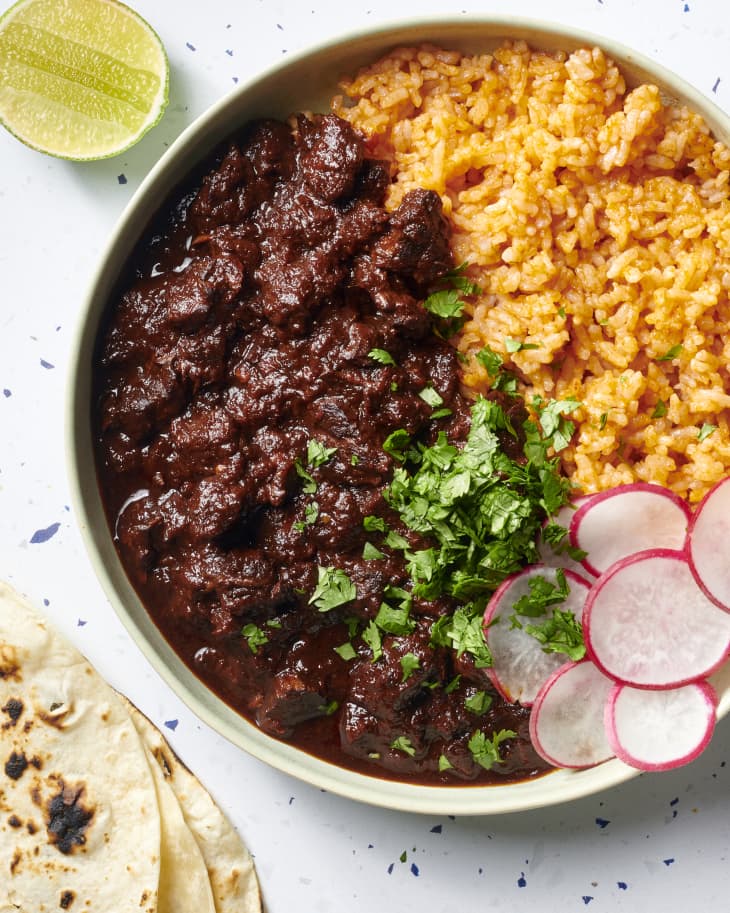 Overhead view of chile colorado and rice in a green bowl topped with radishes and cilantro. Half a lime in the top left corner of the shot, and 3 tortillas peeking in on the bottom left corner of the shot.