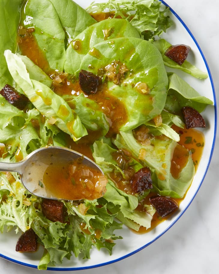 Overhead view of a salad on a white plate with a blue border, being topped with vinaigrette on a spoon.