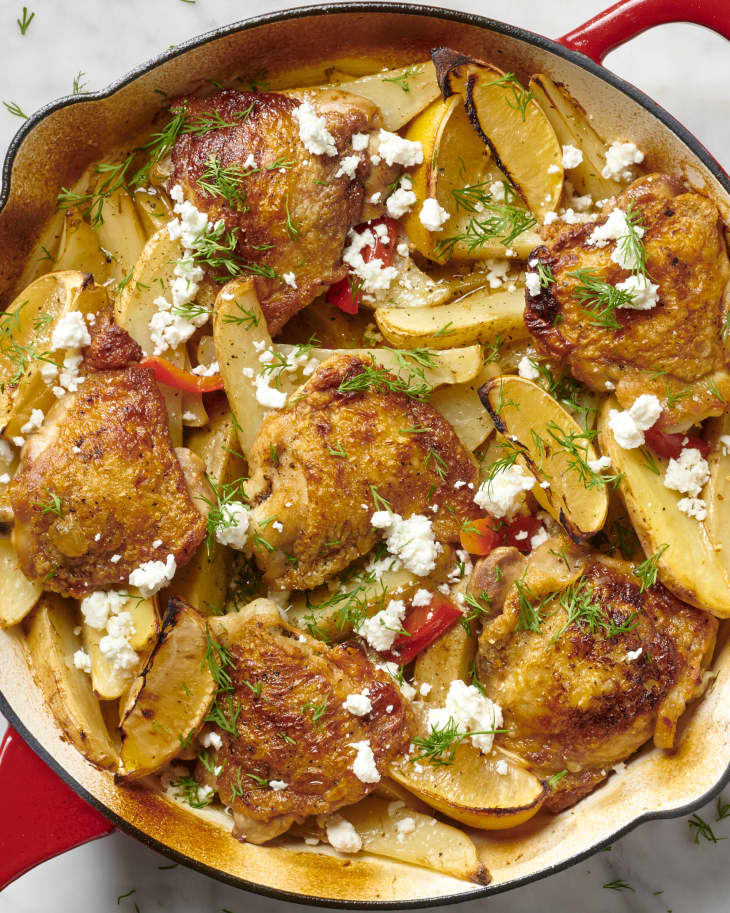 Zoomed in overhead view of chicken and potatoes in red skillet, topped with feta and herbs.
