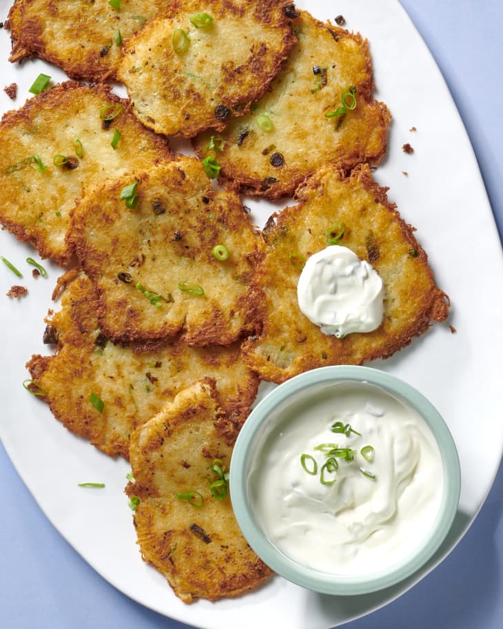 Overhead view of potato pancakes on a white platter topped with herbs and a small blue bowl with sour cream on the side. One potato pancake next to the bowl has a dollop of sour cream on top.