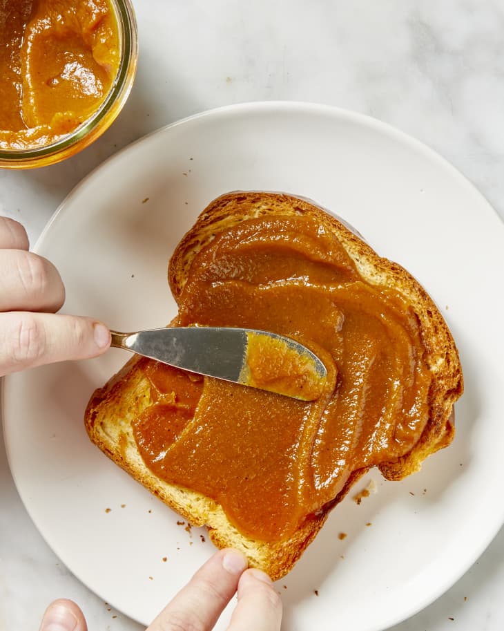 Overhead view of pumpkin butter being spread on a piece of toast, on a white plate, with a jar of pumpkin butter in the top left corner.