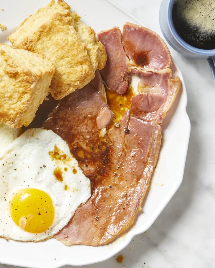 Overhead view of a fried egg, biscuits and ham covered in gravy on a white plate, and a blue mug of coffee in the top right corner.