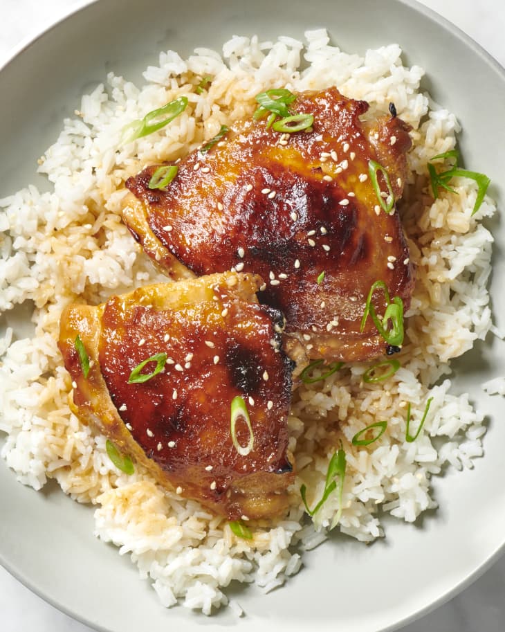 Close up view of two pieces of chicken on a bed of white rice in a light grey bowl, topped with scallions and sesame seeds.