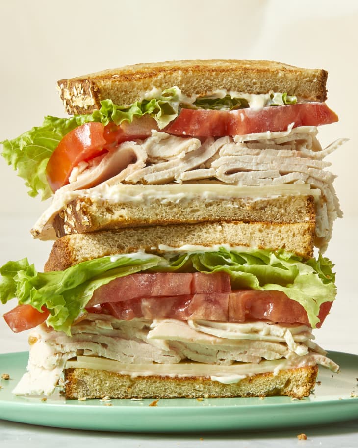 Head on view of a stacked turkey sandwich with cheese, turkey, tomato and lettuce on white bread and a blue plate.