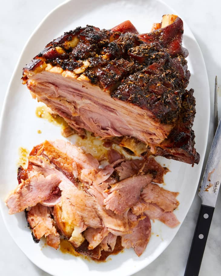 A half-carved baked ham on a serving platter with a carving fork and knife resting next to the platter.