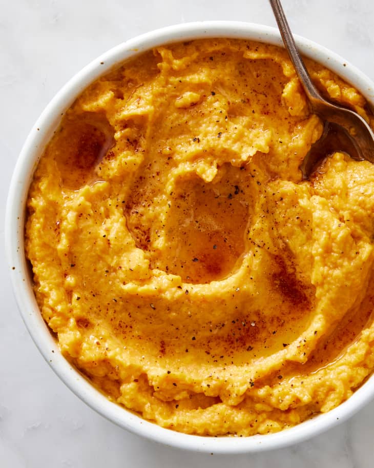 A bowl of rich, orange-toned mashed sweet potatoes with a serving spoon on a marble surface.