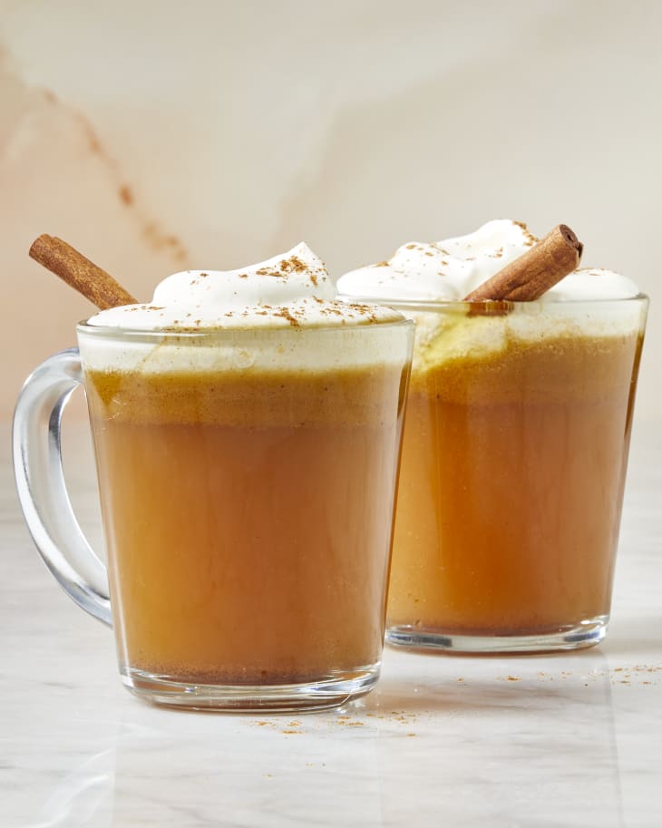 Head on view of two glass mugs of buttered apple cider topped with whipped cream and cinnamon sticks resting in the cups.