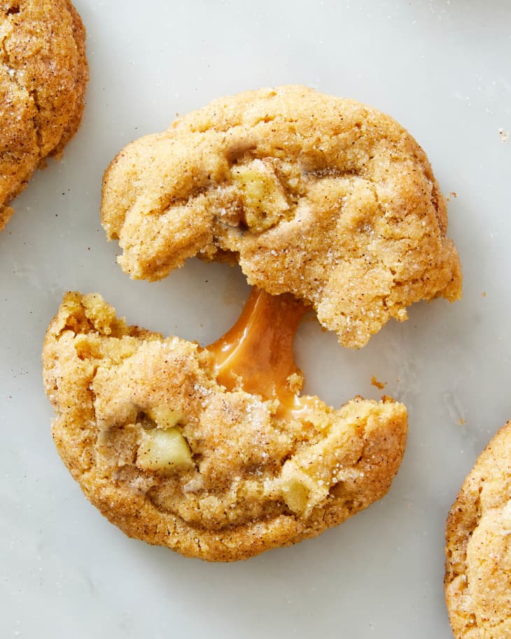 A caramel apple cookie on a marble surface pulled apart in half with gooey caramel connecting the two halves of the cookie.
