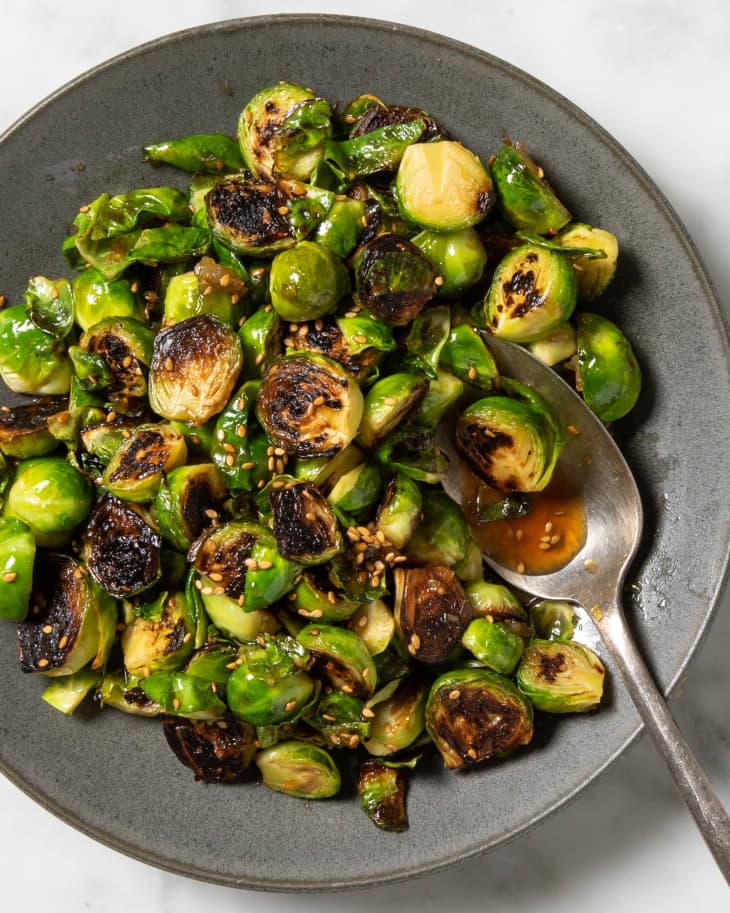 Overhead view of cooked brussels sprouts on a charcoal plate, topped with sesame seeds, and a spoon resting on the plate.