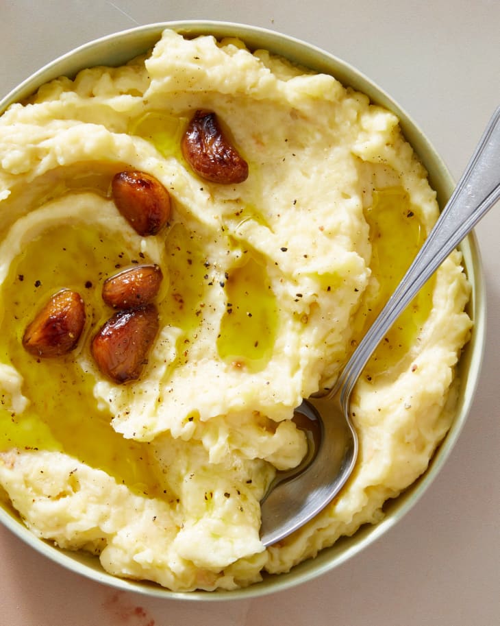 A large serving bowl full of garlic confit mashed potatoes topped with confit garlic with a spoon inserted into the potatoes.