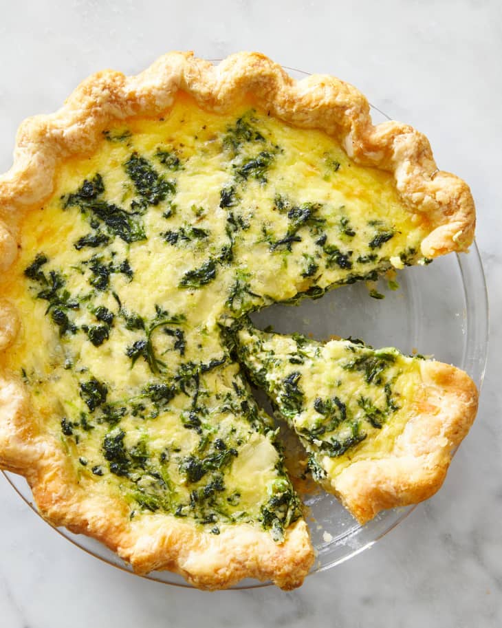 A spinach quiche in a glass pie dish with one slice taken out and another separated from the rest of the quiche.