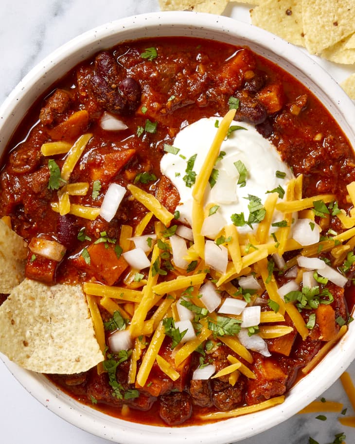 Overhead view of chili in a white bowl, topped with onions, sour cream and cheddar cheese and tortilla chips on the surface to the right of the bowl.