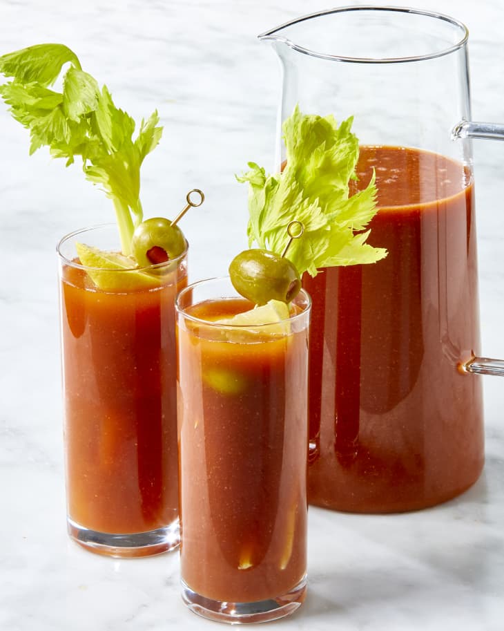 Head on shot of a pitcher and two glasses of bloody mary's, garnished with lemon, olives and celery.