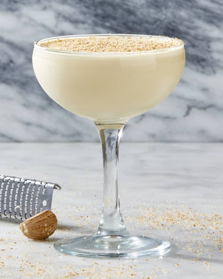 Head on shot of a coupe glass of brandy alexander, on a white marble surface, and nutmeg shavings around it.