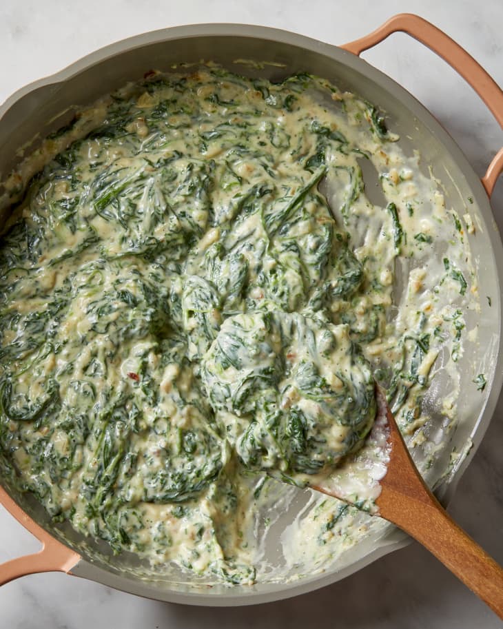 Overhead shot of creamed spinach in a light pink colored pan, with a wood spoon scooping it out.