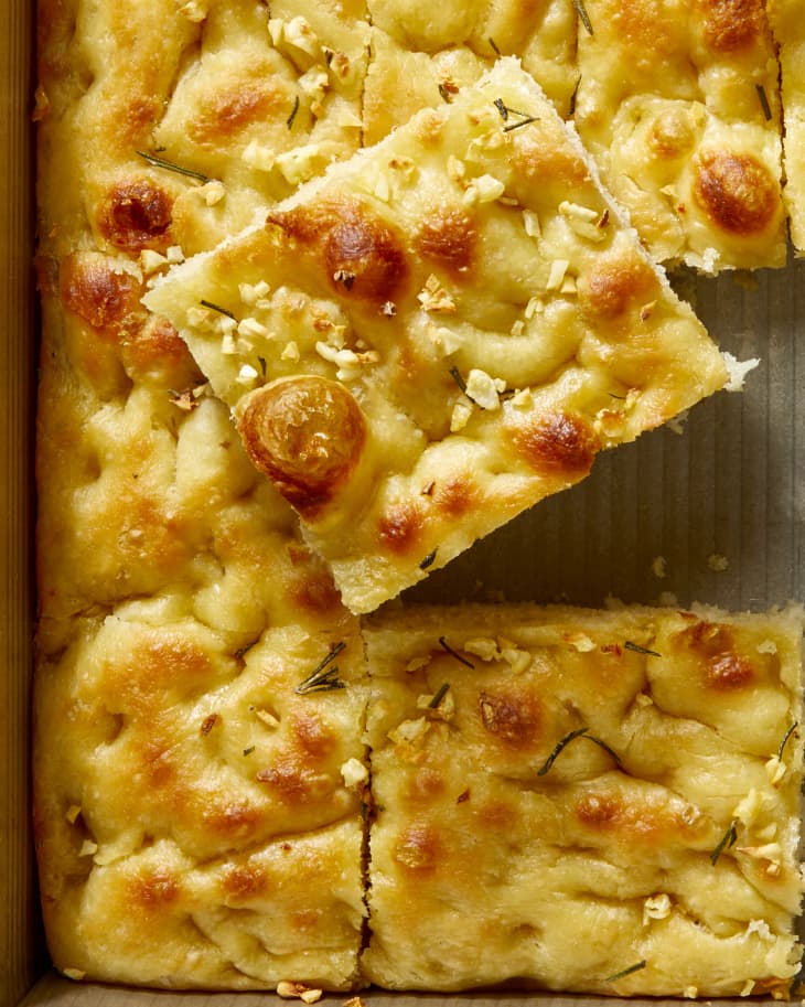 Close up of baked focaccia topped with herbs and garlic, with one piece cut out and stacked on the loaf.