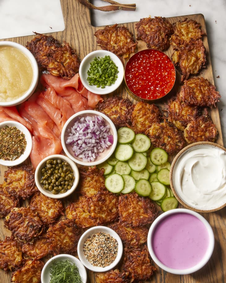 Overhead shot of a large wooden cutting board with filled with latkes and an assortment of toppings in small white bowls.