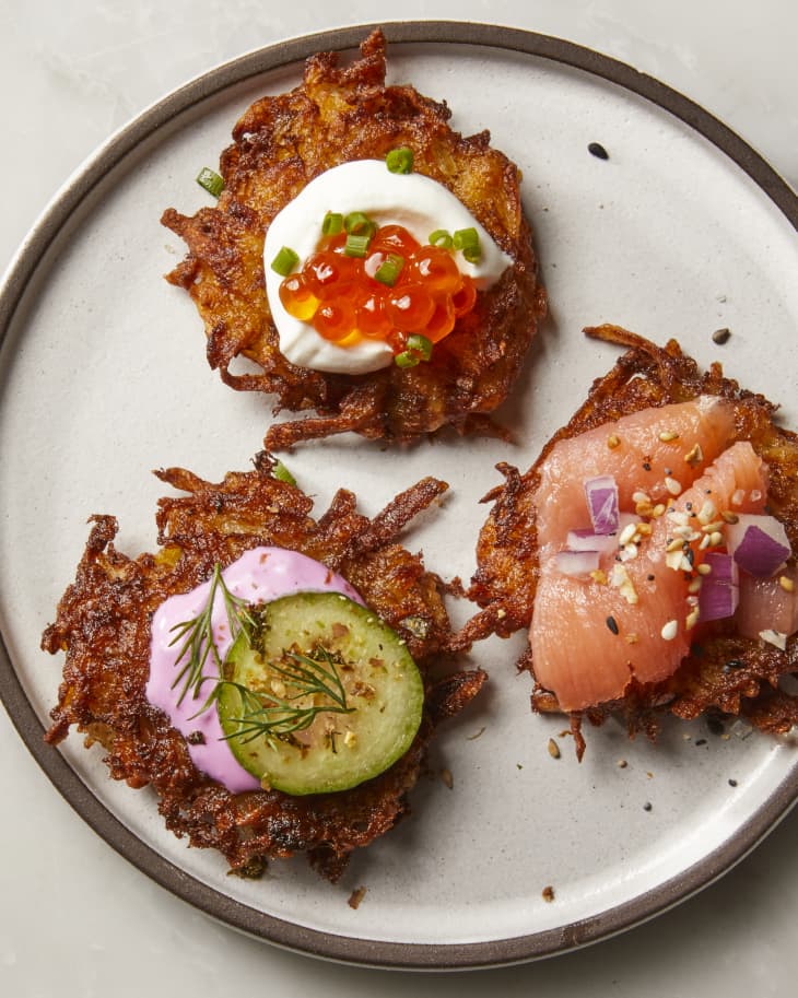 Overhead shot of three latkes on a grey plate with a brown rim.  The top latke is topped with sour cream, roe and green onions, the latke to the right is topped with smoked salmon and red onion and the latke on the left is topped with a pickle and dill.