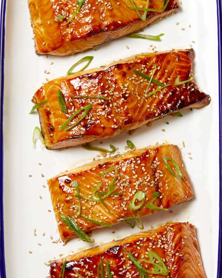 Overhead shot of four pieces of teriyaki salmon on white surface, topped with scallions and sesame seeds.