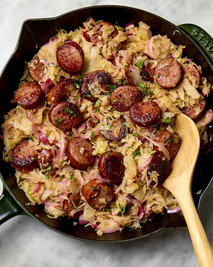 Overhead shot of cooked sauerkraut and sausage in a cast iron pan, with a wooden spoon resting in the bottom right part of the pan.