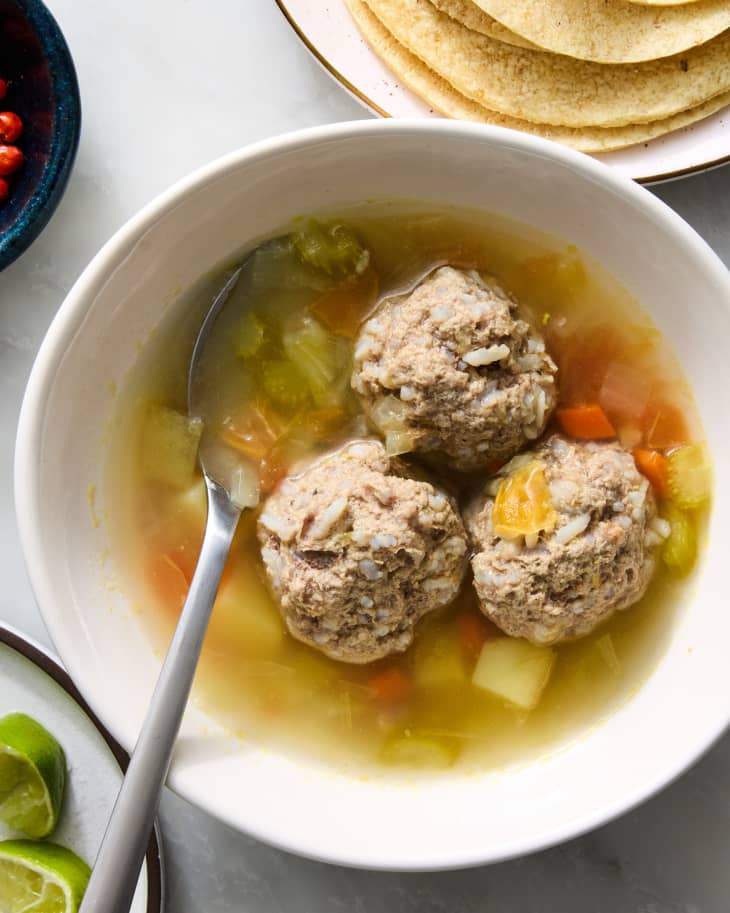 Overhead shot of albondigas soup with three meatballs in a small white bowl, with a metal spoon resting in the bowl.