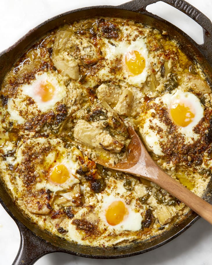 Overhead shot of spinach artichoke baked eggs in a cast iron pan, with a wooden spoon resting with a spoonful on it in the middle of the pan.