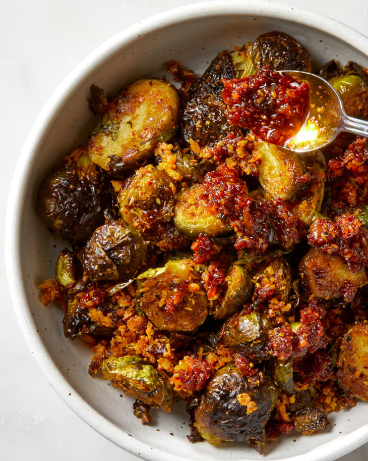Overhead shot of chili crisp brussels sprouts in a white bowl on a white marble surface, with a spoonful of chili crisp being drizzled on top.
