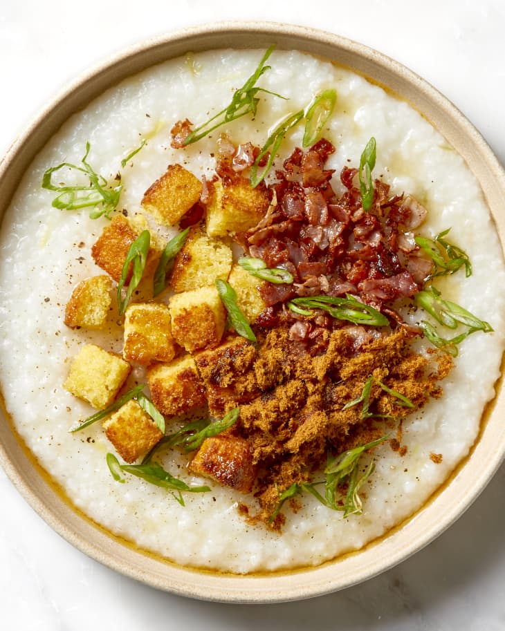 Overhead shot of congee in a shallow light brown bowl, topped with pork floss, bacon, cornbread pieces and green onion.