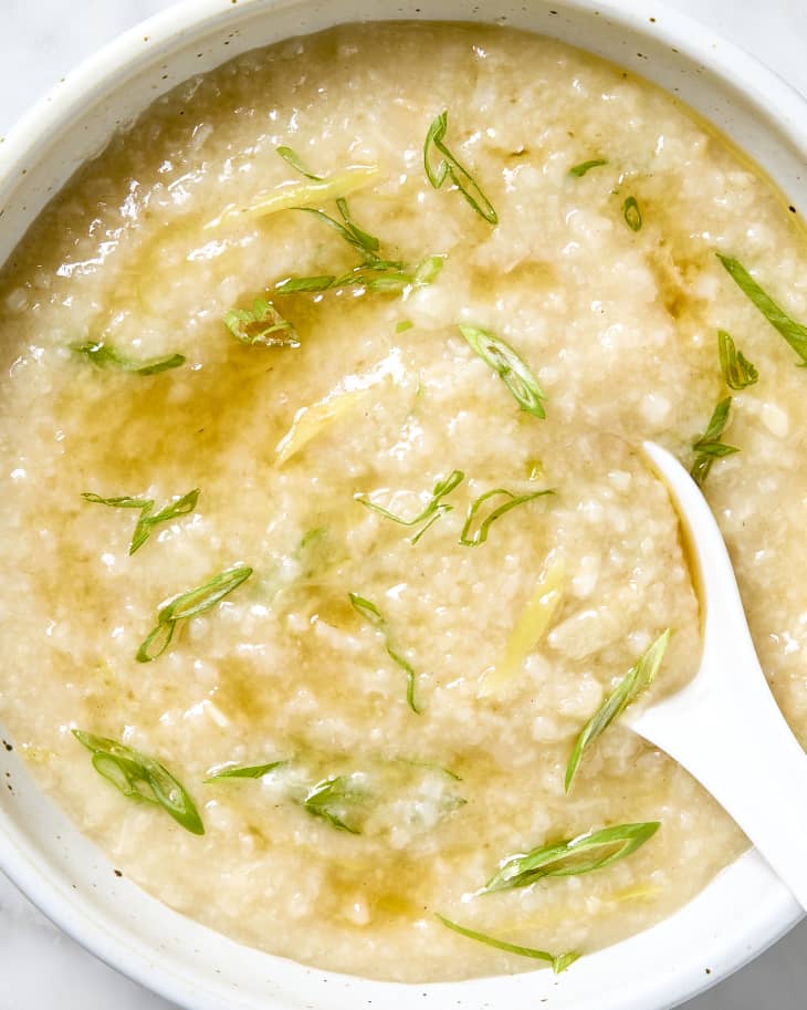 Overhead shot of congee in a white bowl, topped with green onion and theres a white soup spoon resting on the right side of the bowl.