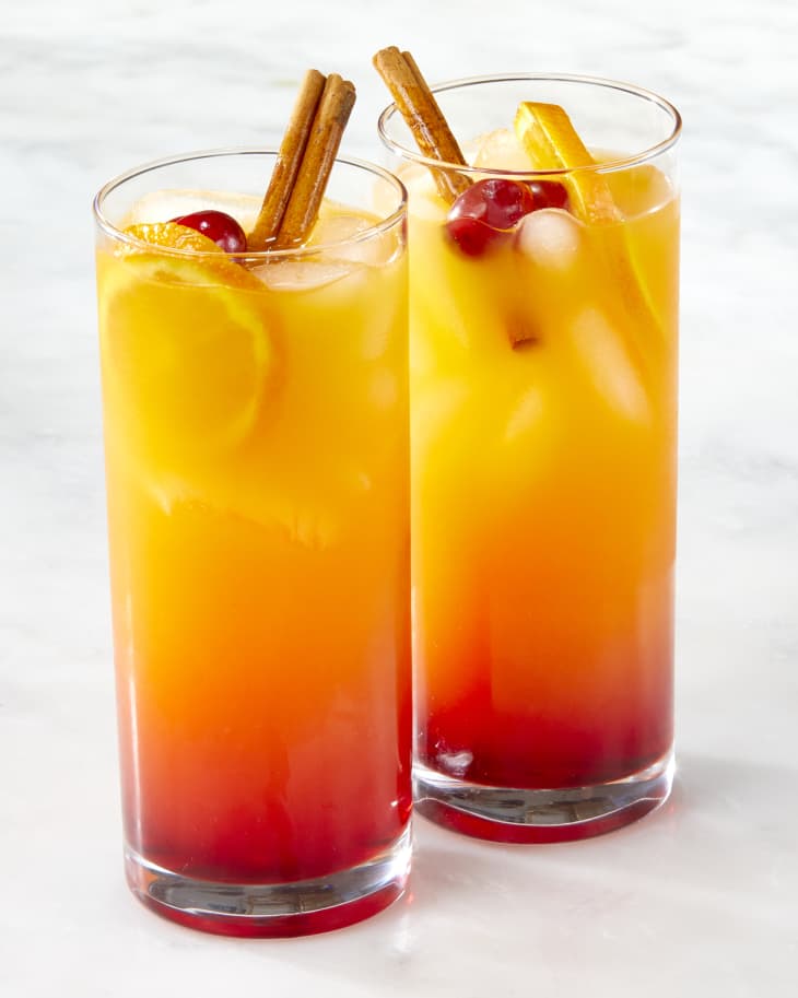 Head on shot of two cranberry tequila sunrises in collins glasses, and garnished with an orange wheel, cinnamon stick and maraschino cherries.